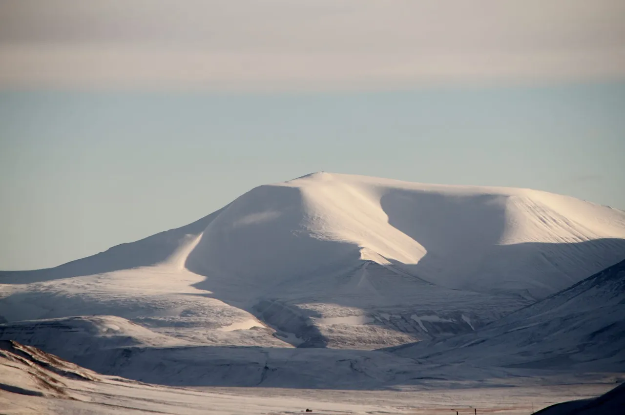 A broad, snow-covered mountain with a rounded summit rising above a snowy valley in soft evening light