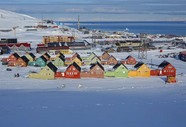 A row of brightly painted wooden houses in red, yellow, green and blue on the snow in Longyearbyen, with the fjord behind