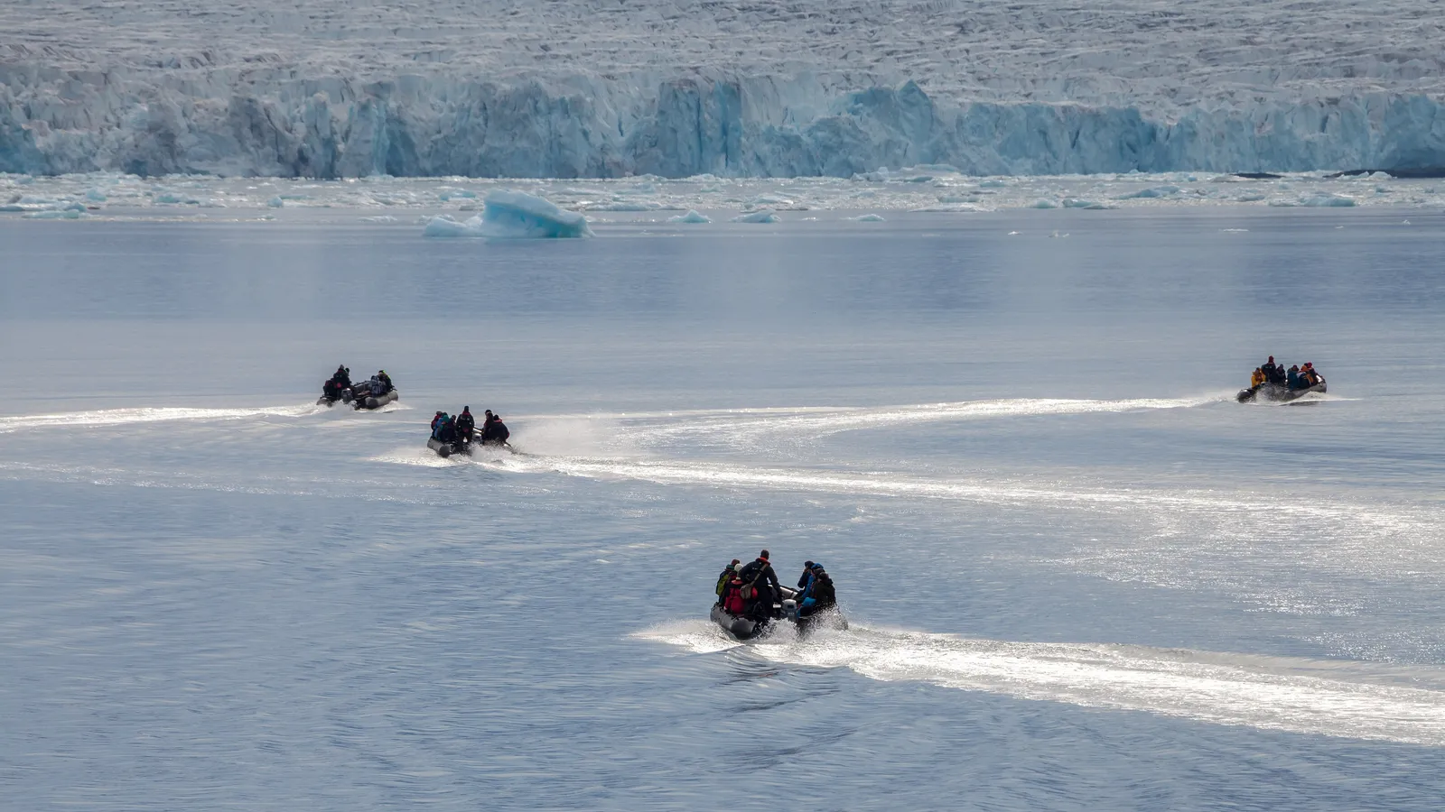 Four small inflatable boats full of passengers leave white wakes across a calm fjord towards a glacier front scattered with ice