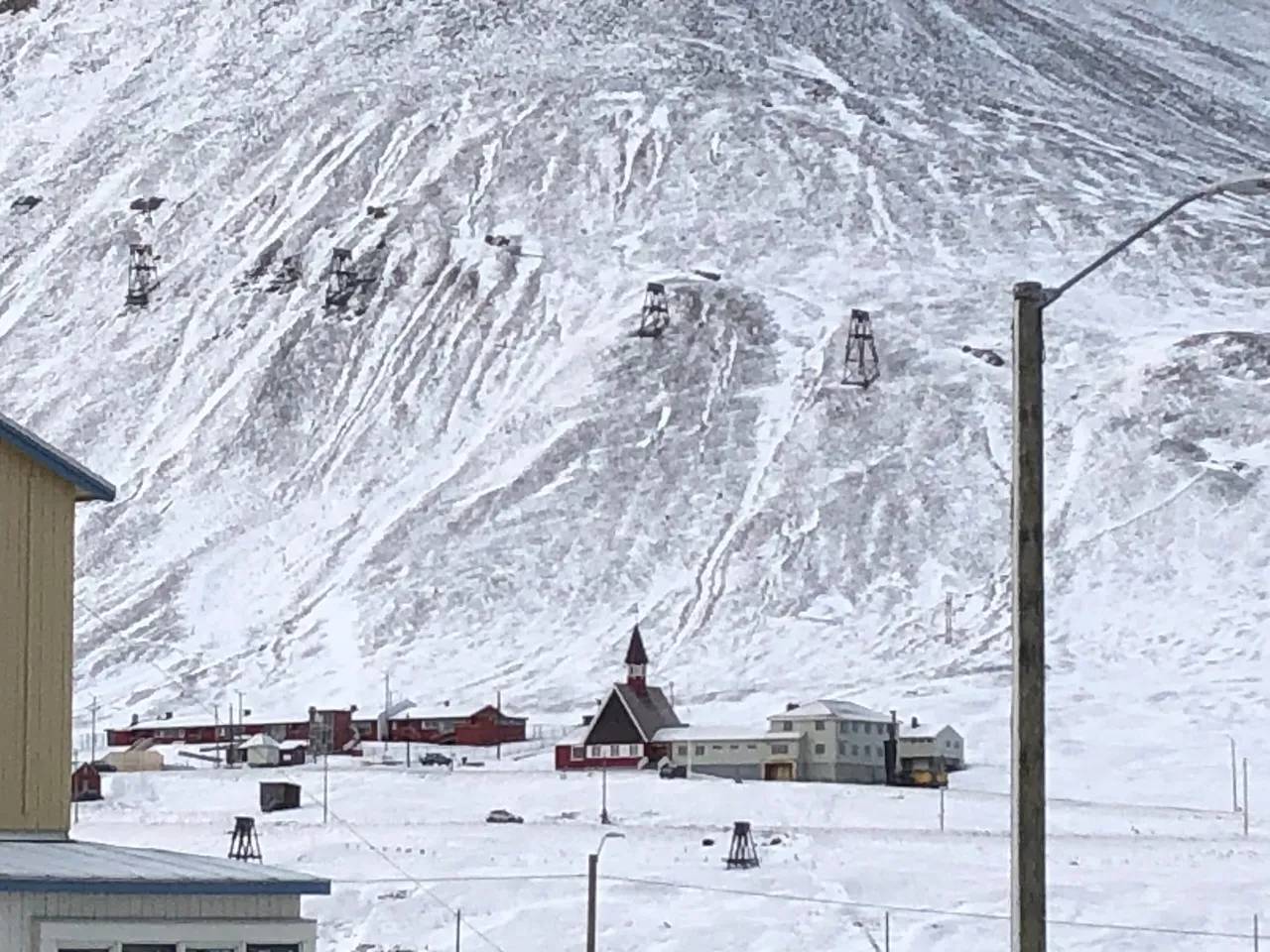 Svalbard Church, a dark red wooden church with a pointed tower, among houses below a snowy mountainside lined with old mining cableway towers