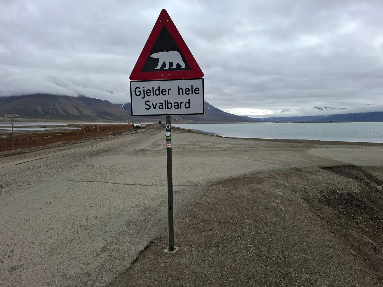 Ein dreieckiges Verkehrsschild mit rotem Rand, das einen Eisbären und die Worte „Gjelder hele Svalbard“ zeigt, neben einer Schotterstraße am Fjord