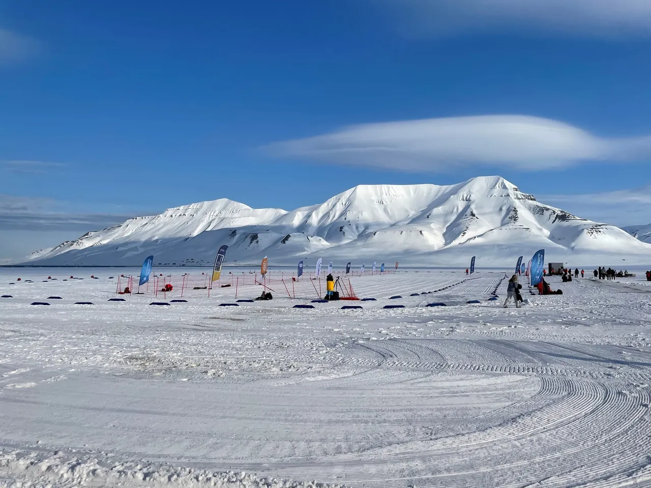 Eine weite, präparierte Schneearena mit Fahnen und Zelten vor schneebedeckten Bergen unter blauem Himmel mit linsenförmigen Wolken