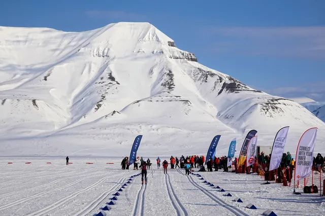 Skiers crossing the finish of the Svalbard Ski Marathon in a snowy valley, with sponsor flags lining the tracks and a big snow-covered mountain behind