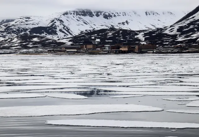 隔着漂浮的海冰远眺皮拉米登的砖砌建筑，后方是带着积雪条纹的深色山峦
