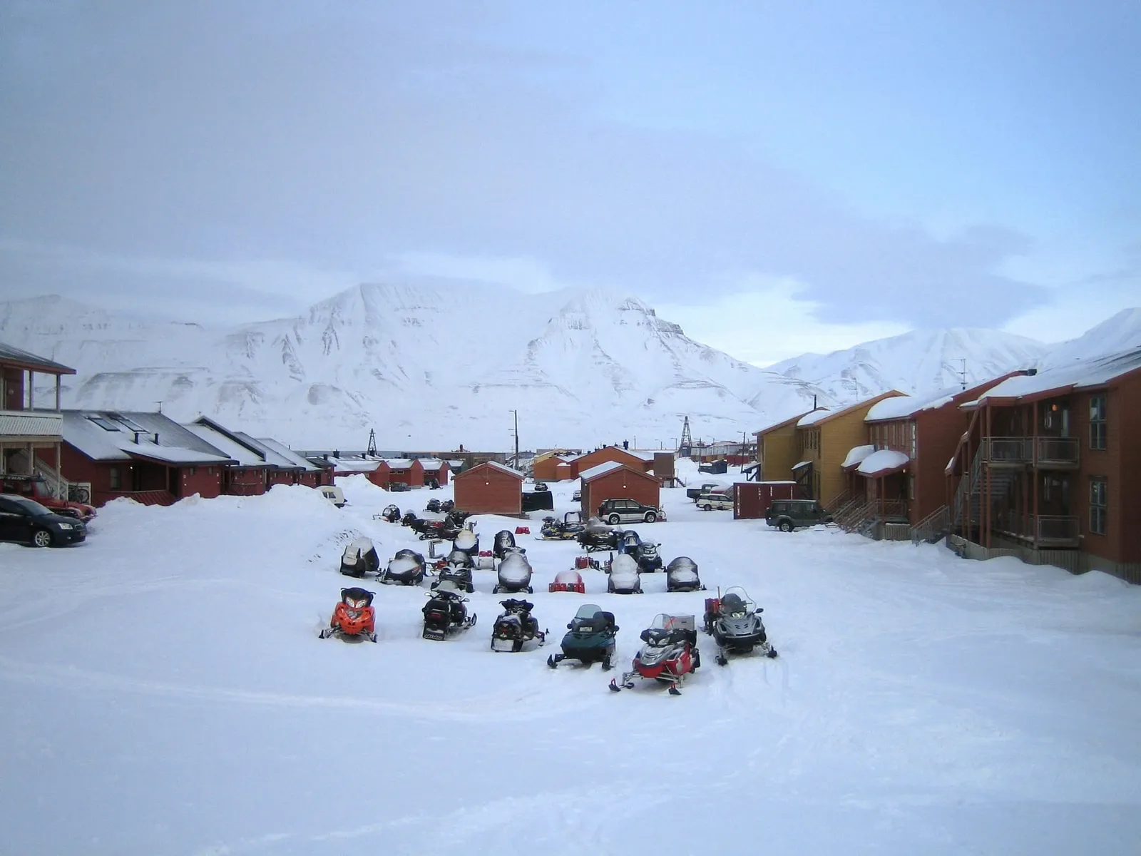Dutzende Schneemobile, in Reihen auf dem Schnee zwischen Holzhäusern in Longyearbyen abgestellt, dahinter weiße Berge