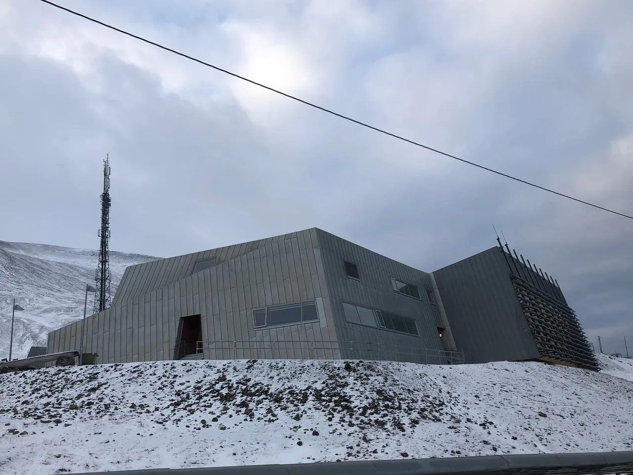 Un edificio gris moderno de paredes angulosas y un alto mástil de radio, en una ladera nevada bajo un cielo nublado
